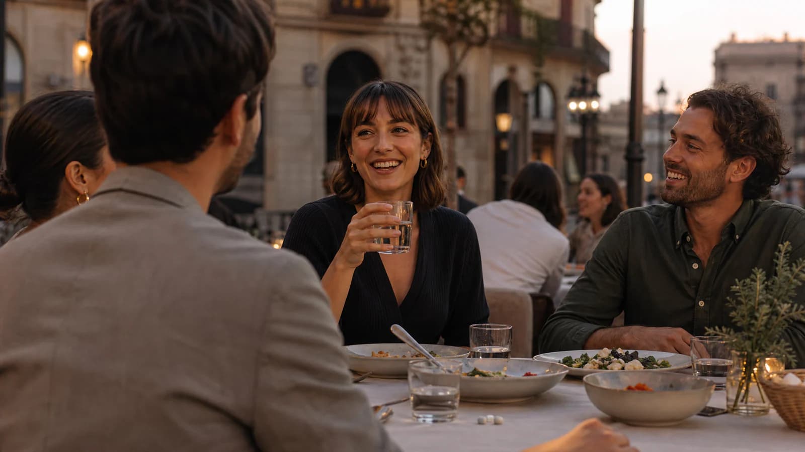 Friends sharing dinner and glasses of water on a European restaurant terrace at dusk, with capsules on the table.