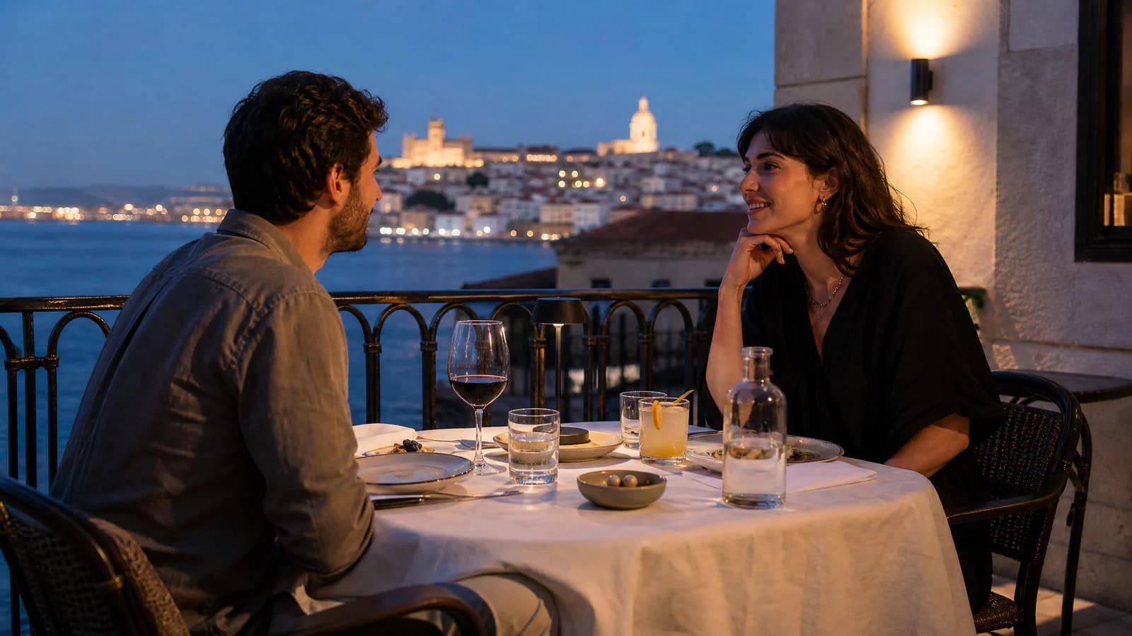 A man and woman sit across from each other at a restaurant table on a waterfront terrace, with drinks and plates before them.